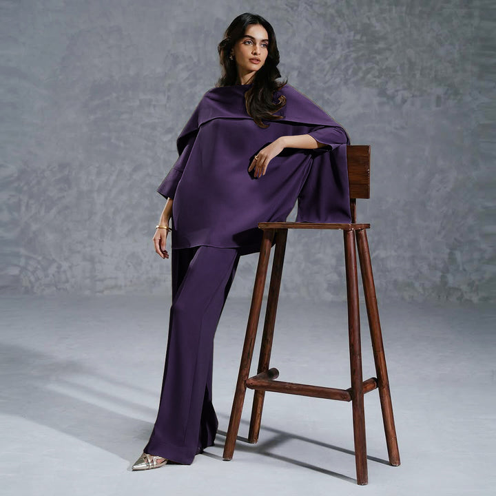 Woman in a purple outfit sitting on a wooden stool against a gray background