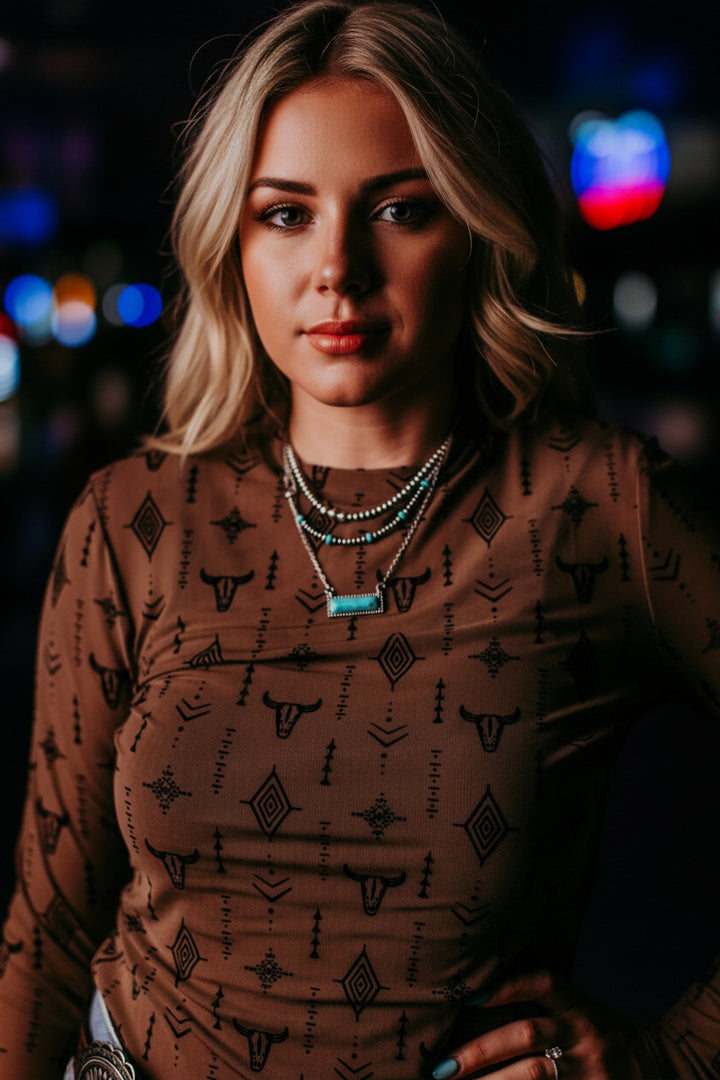 A close-up of a woman wearing a brown long-sleeve top with a black geometric and tribal-inspired pattern, accessorized with layered necklaces and a western belt.