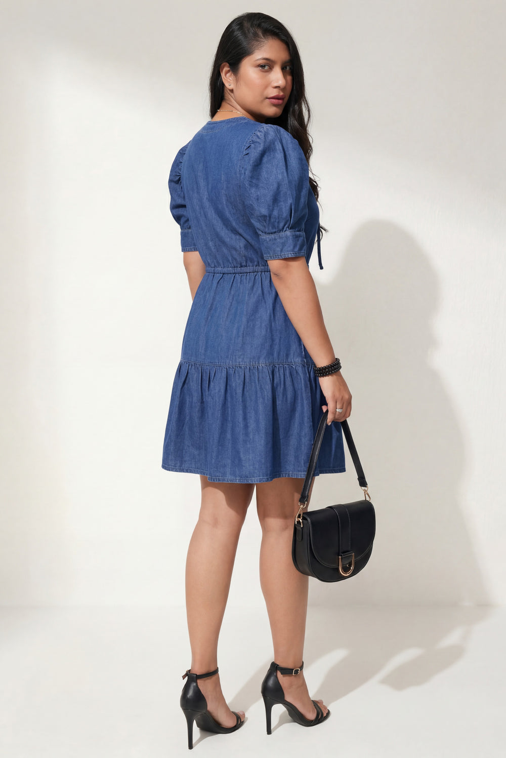Errabelly blue denim mini dress with drawstring waist, puff sleeves, and tiered skirt. Model is wearing black heels and carrying a black handbag.