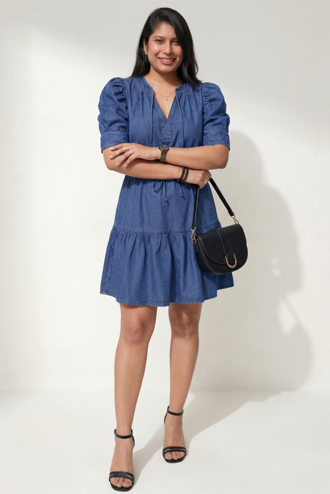 Errabelly blue denim mini dress with puff sleeves, drawstring waist, and tiered skirt. Model is wearing black heels and carrying a black handbag.